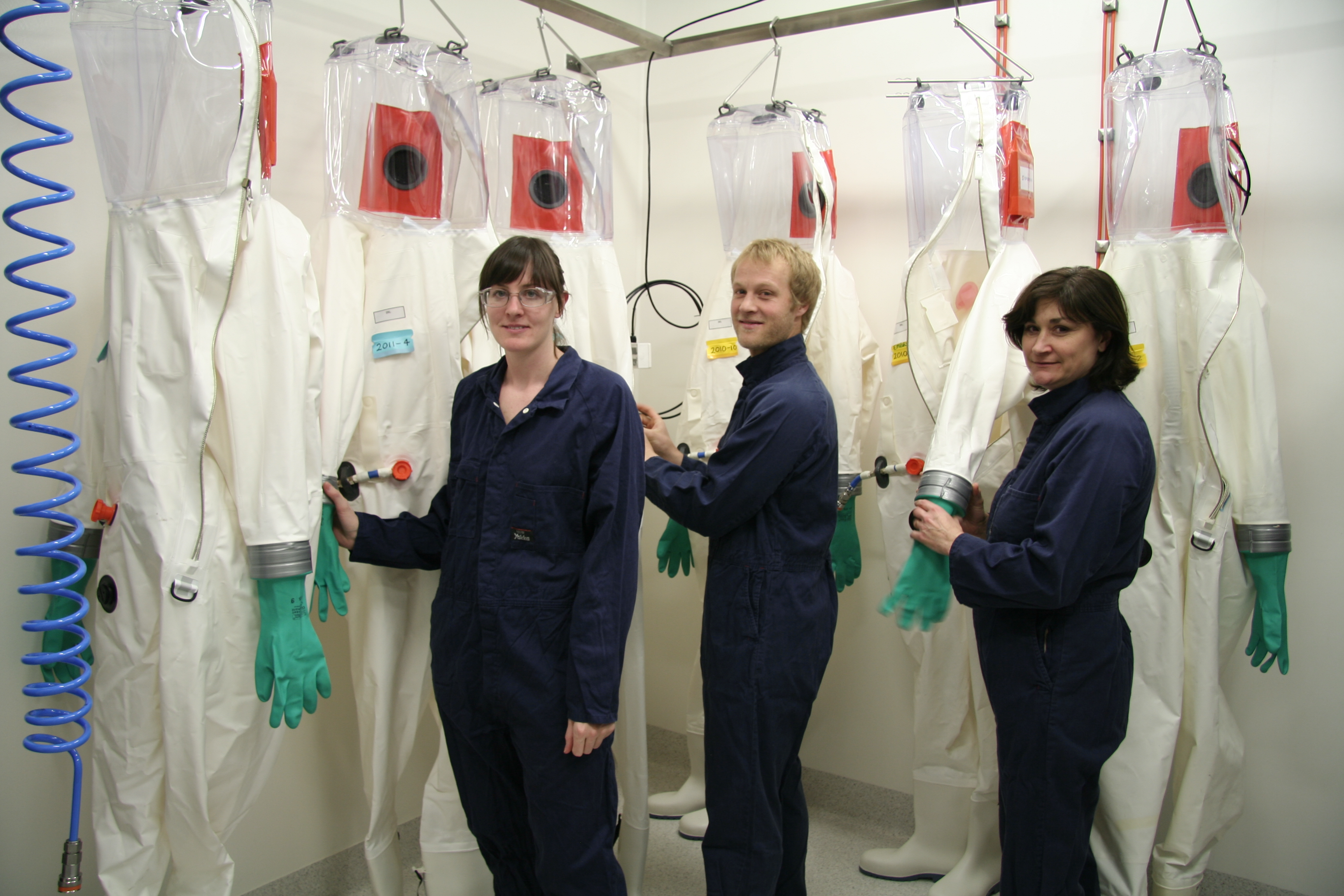 Three people wearing blue overalls standing infront of encapsulated suits that are hanging from hooks