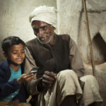 Happy village boy sitting beside his grandfather and playing with his grandfather's mobile phone. They are sitting against a background of an adobe wall of a rural house in India.