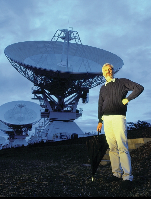 A man with a folded umbrella standing in front of telescope dishes.