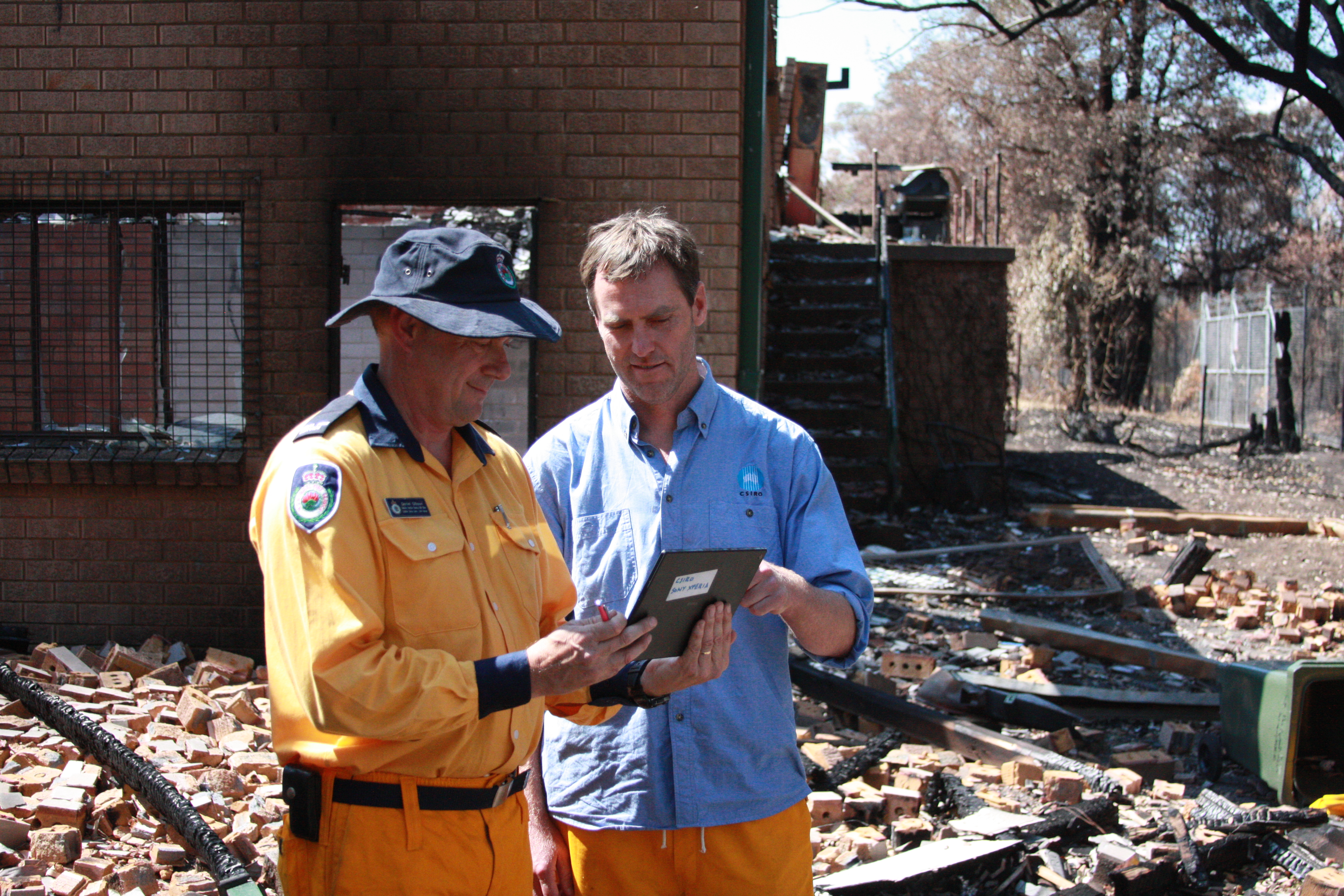 Two men holding a smart tablet standing in front of a burnt house