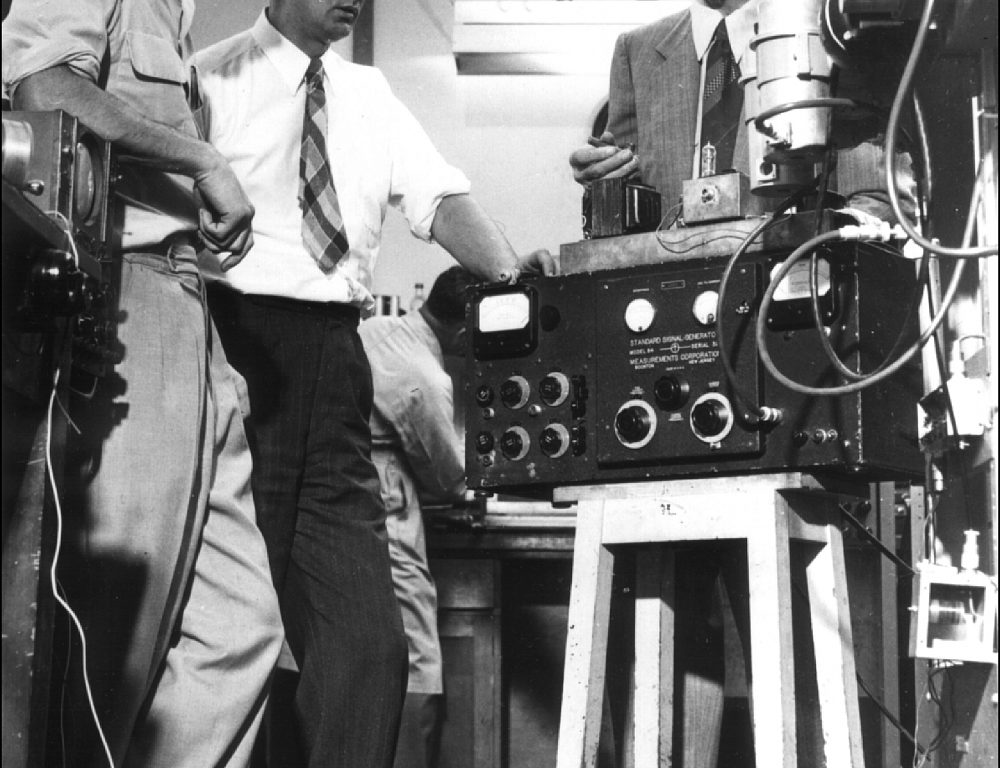 Three men standing near radio equipment in a laboratory in the late 1940s.