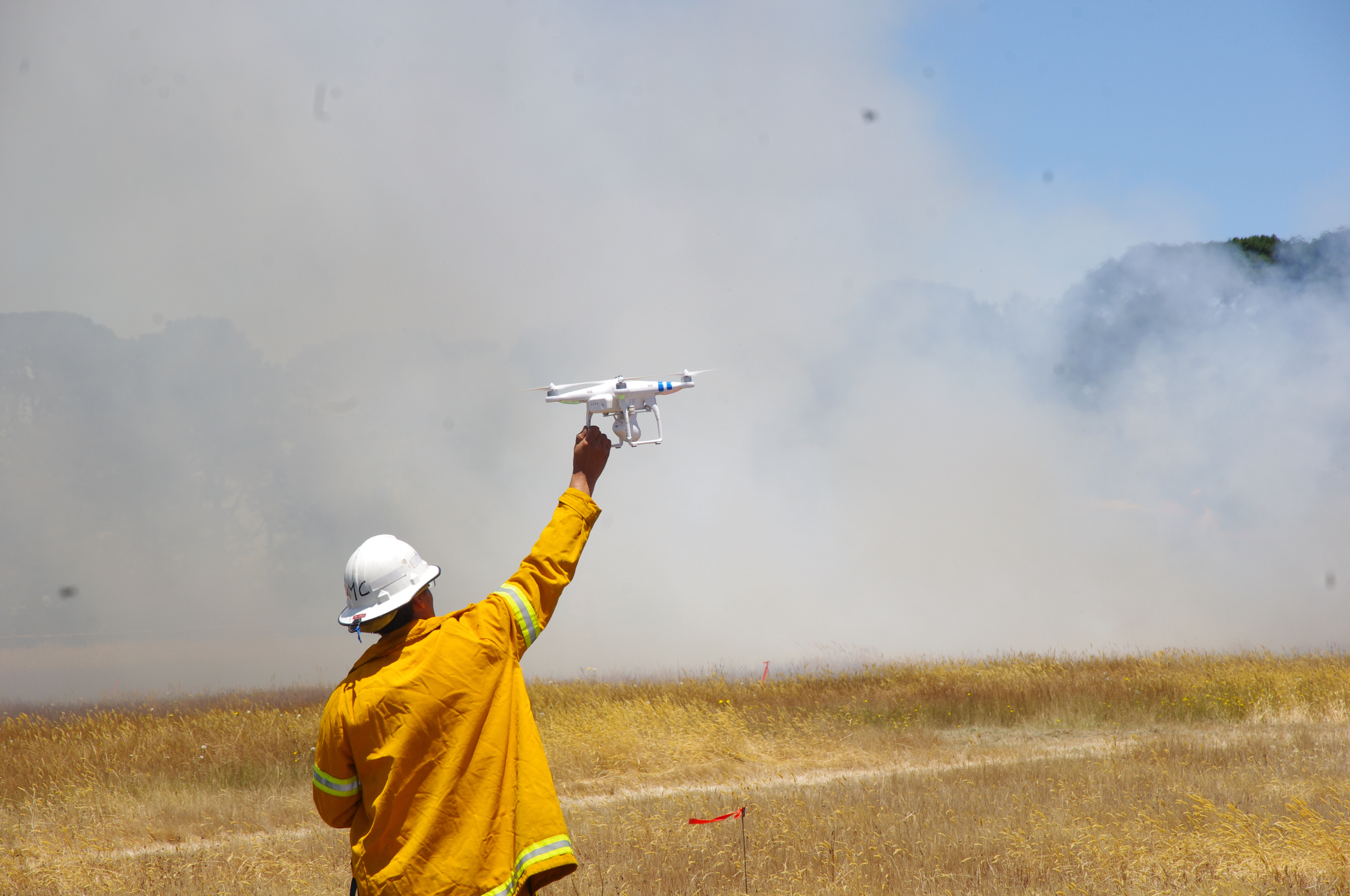 A scientist in fire gear holds up a UAV with smoke in the background
