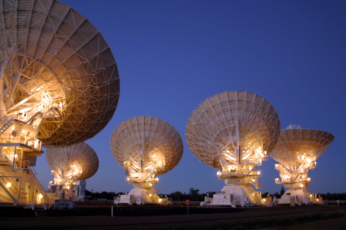 Five white radio telescope dishes point to the night sky, the dishes are illuminated