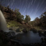 A waterfall over rocks and a river in the foreground and star trails in the night sky at the top of the image.