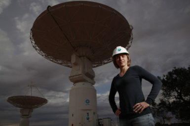 In the foreground a woman stands with her hands on her hips, there are large, while telescope antennas in the background.