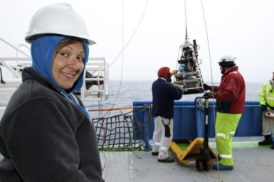 Woman in left of picture wearing jumper and hardhat on boat with scientific equipment hanging from cable background. Ocean, Investigator, Voyages