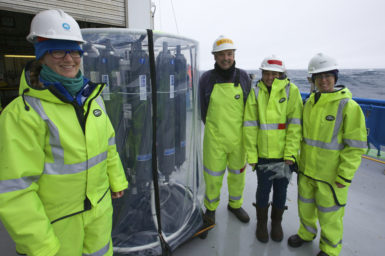 Four people wearing yellow high vis suits and hard hats standing on the deck of a ship with piece of marine sampling equipment