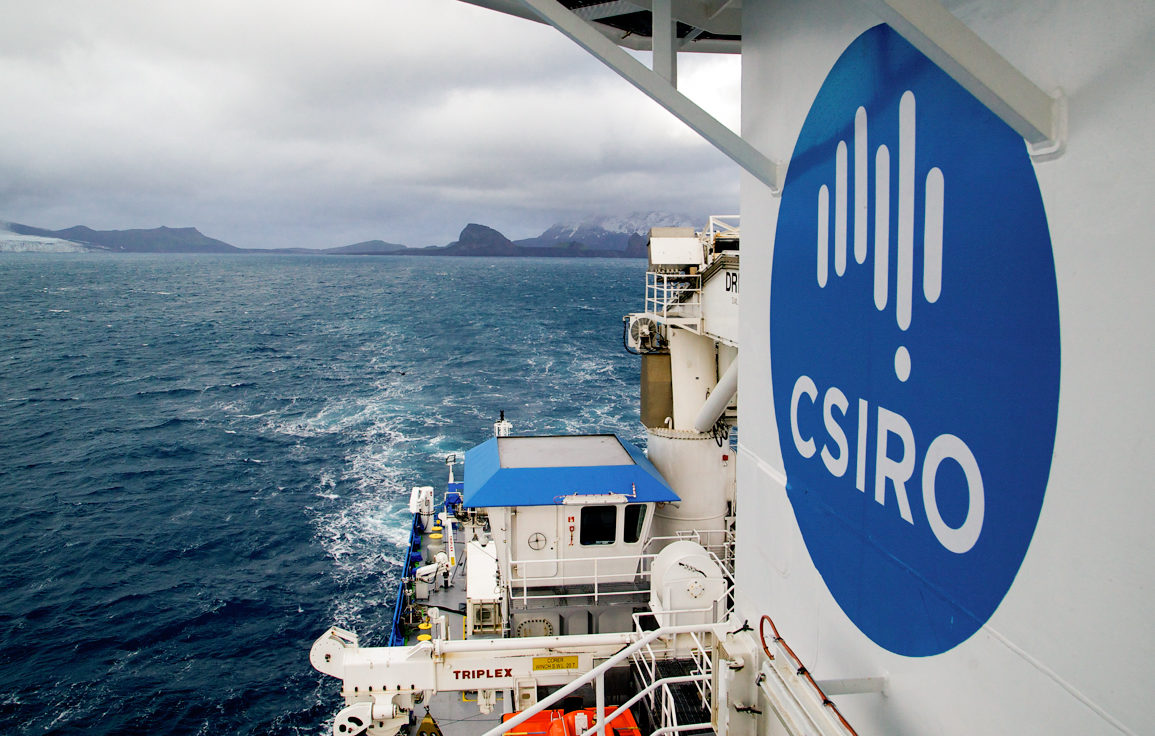 View from top of ship with islands in background and large circualr blue and white CSIRO logo in foreground