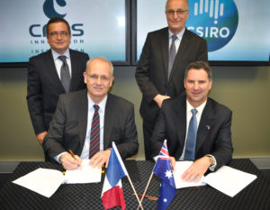 Four men in dark suits, two standing, two sitting signing agreemnet, French and Australian flags in the foreground on the table.