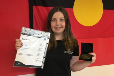 Sharni Cox standing in front of the Australian Aboriginal Flag with the medal and certificate for the 2016 Student STEM Award.