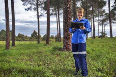 Female in blue overalls staining in a field holding a tablet device