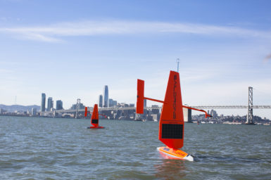 Two Saildrones seen sailing in the water. Buildings are visible in the far distance.