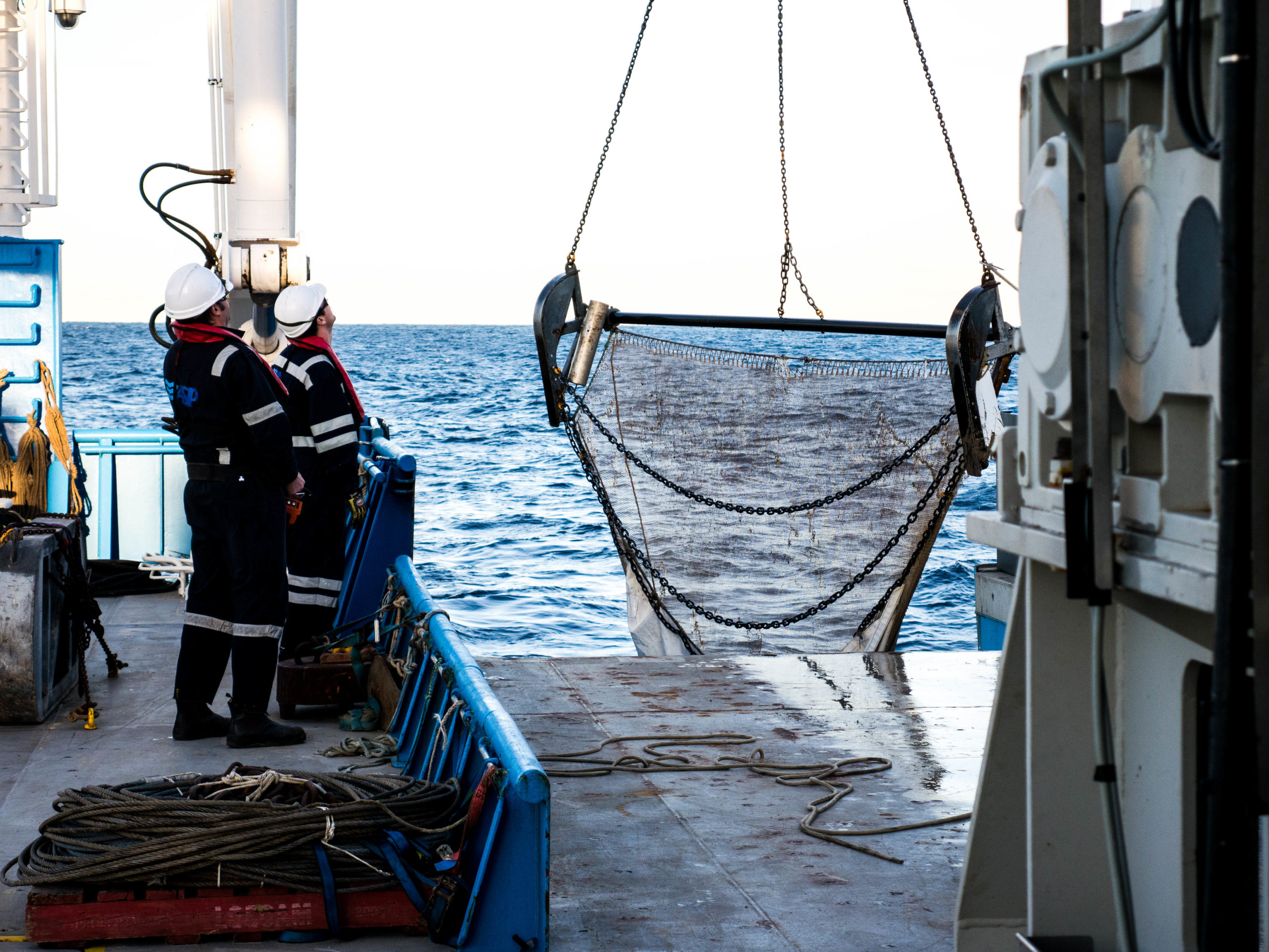Two people aboard a ship watching a large net being hauled back onto the deck.