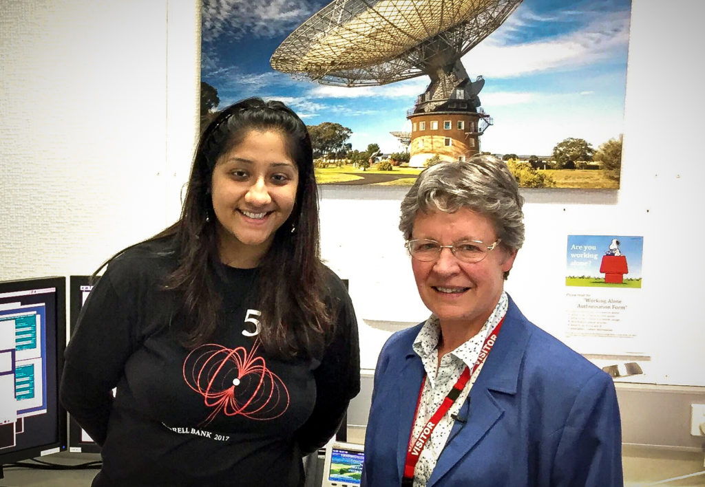 Two women standing next to each other looking at camera with picture of radio telescope on wall in background.