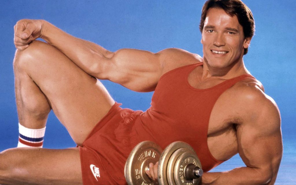 A 1980s photo of Arnold Schwarzenegger smiling at the camera holding weights
