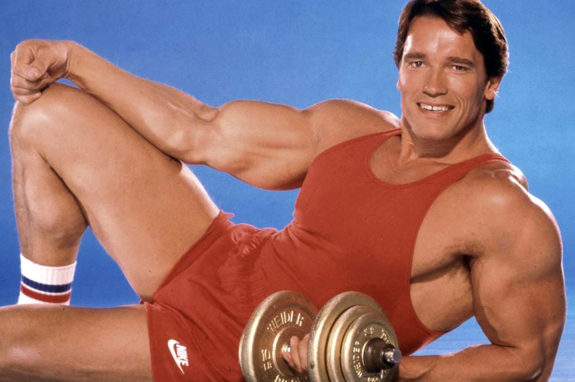 A 1980s photo of Arnold Schwarzenegger smiling at the camera holding weights