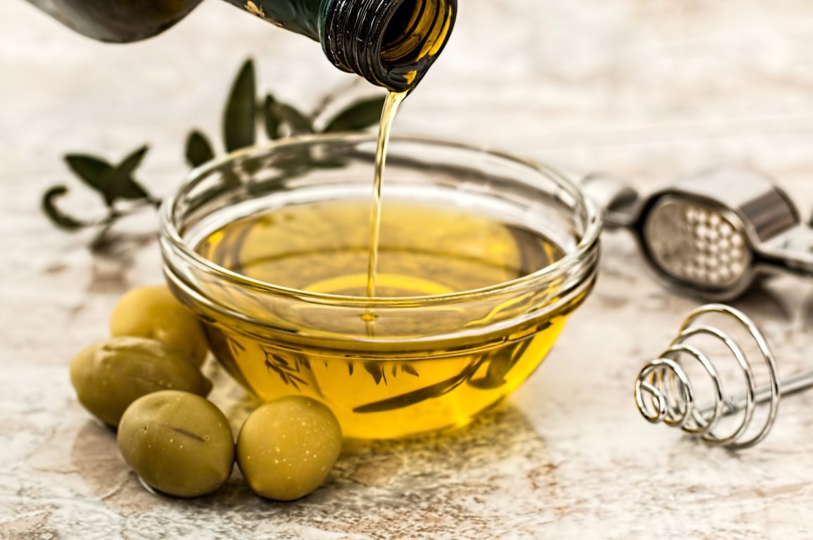 Olive oil being poured from a bottle into a small dish with fresh olives alongside the dish