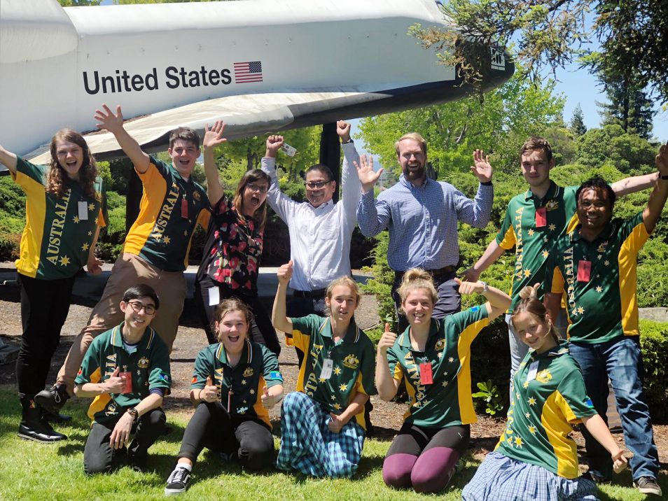 A group of science students and teachers under a plane marked with USA flag. 