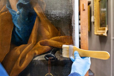 art resin is applied to a painting with a wide brush by a gloved hand