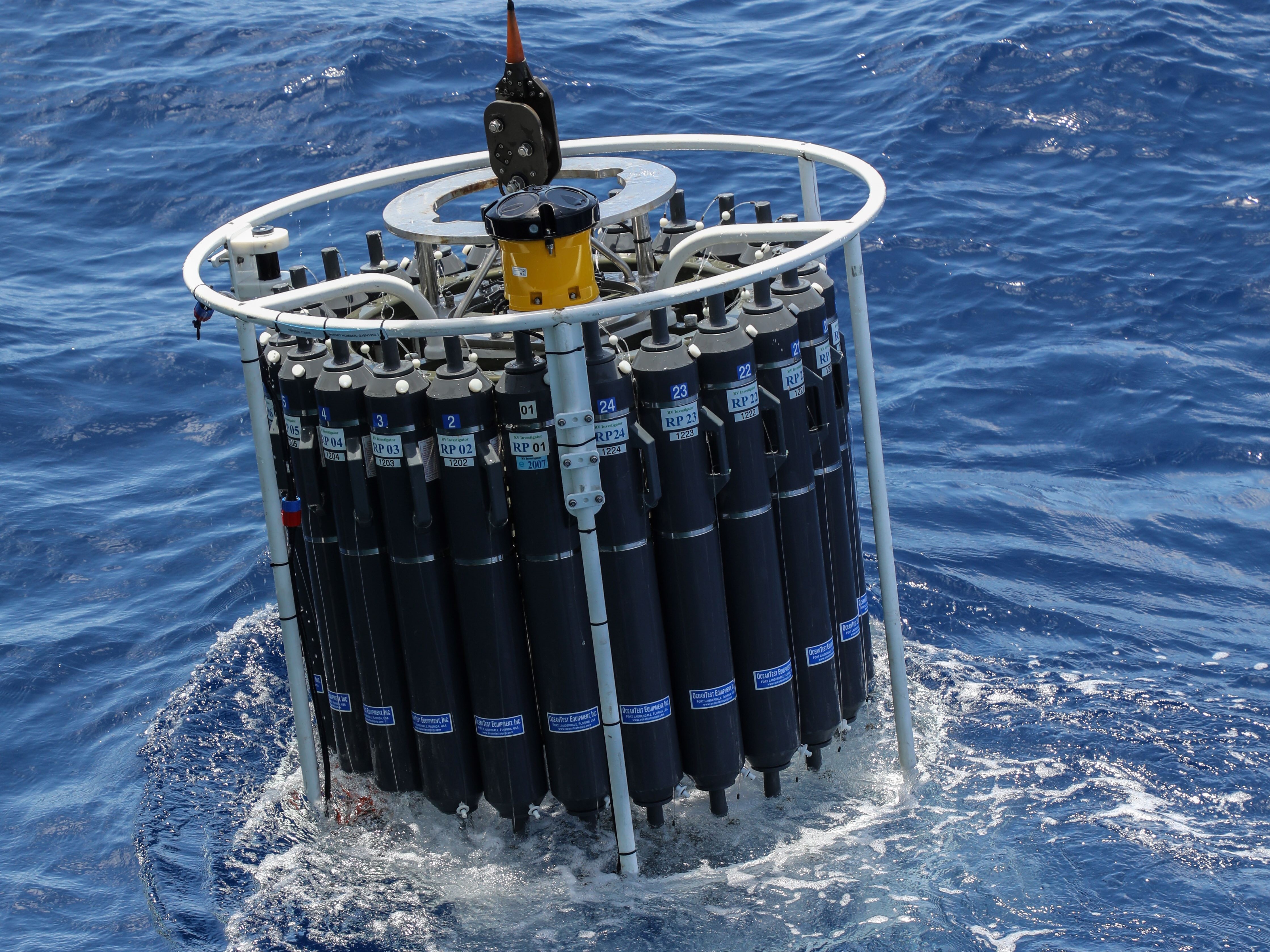 A large cylinder filled with black bottles is lowered into the ocean
