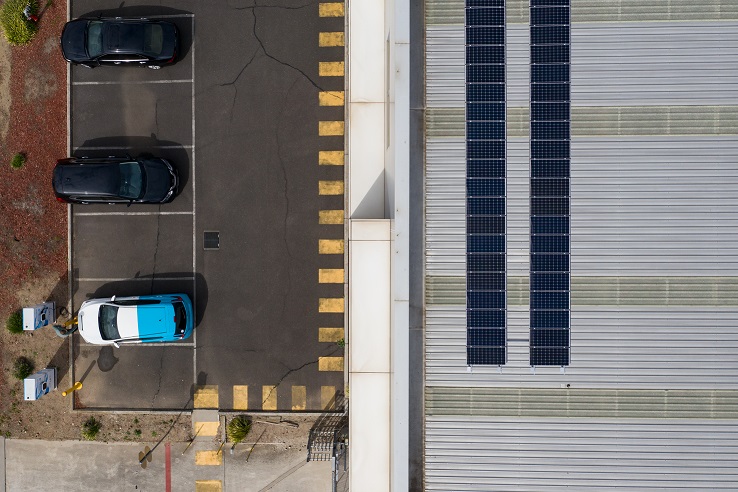 drone shot of a carpark on the left half of the image and then a solar panel covered building on the right. There is a column of cars, two at the top are black and the bright coloured blue and white car is an electric vehicle being charged. 