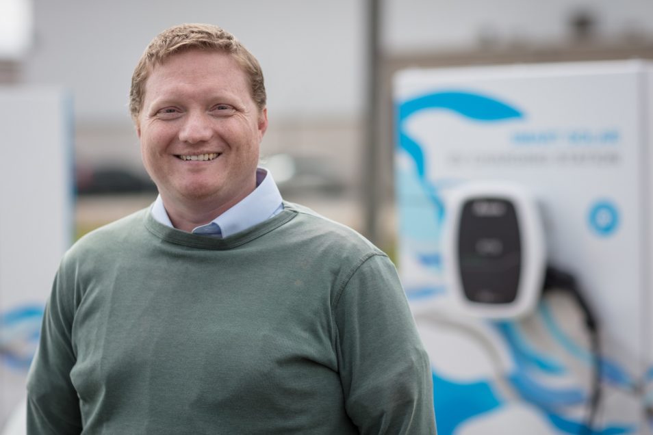 man smiling with a greeny-grey jumper on. The charging station is not in focus but it's in the background