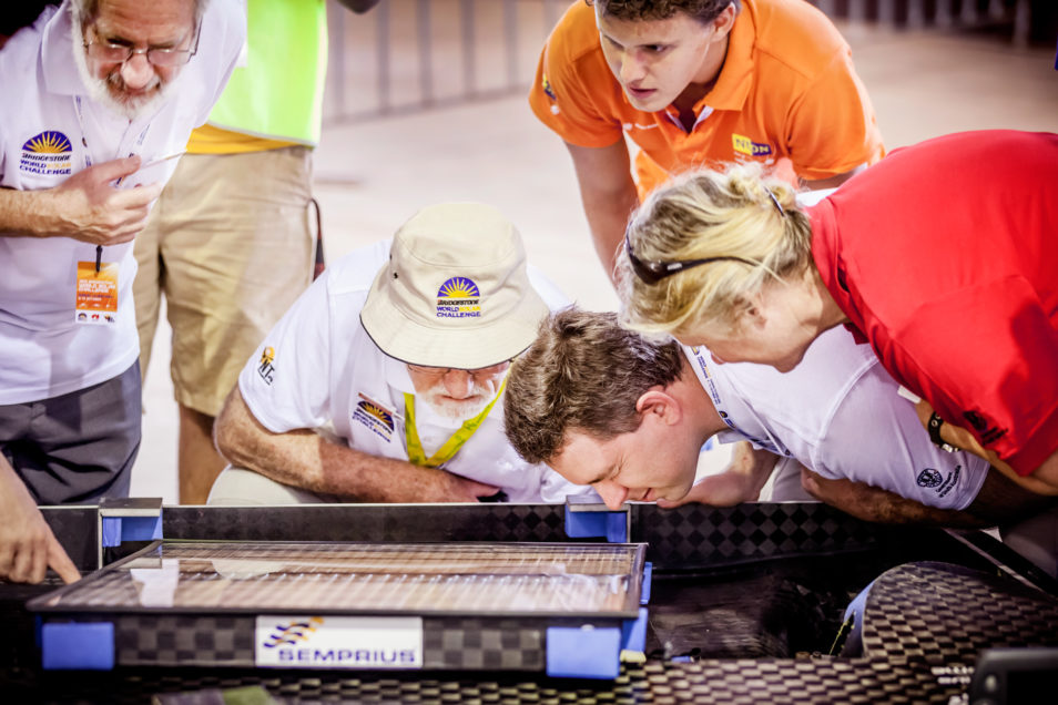 A group of people peering down and examining a tray-looking battery. 