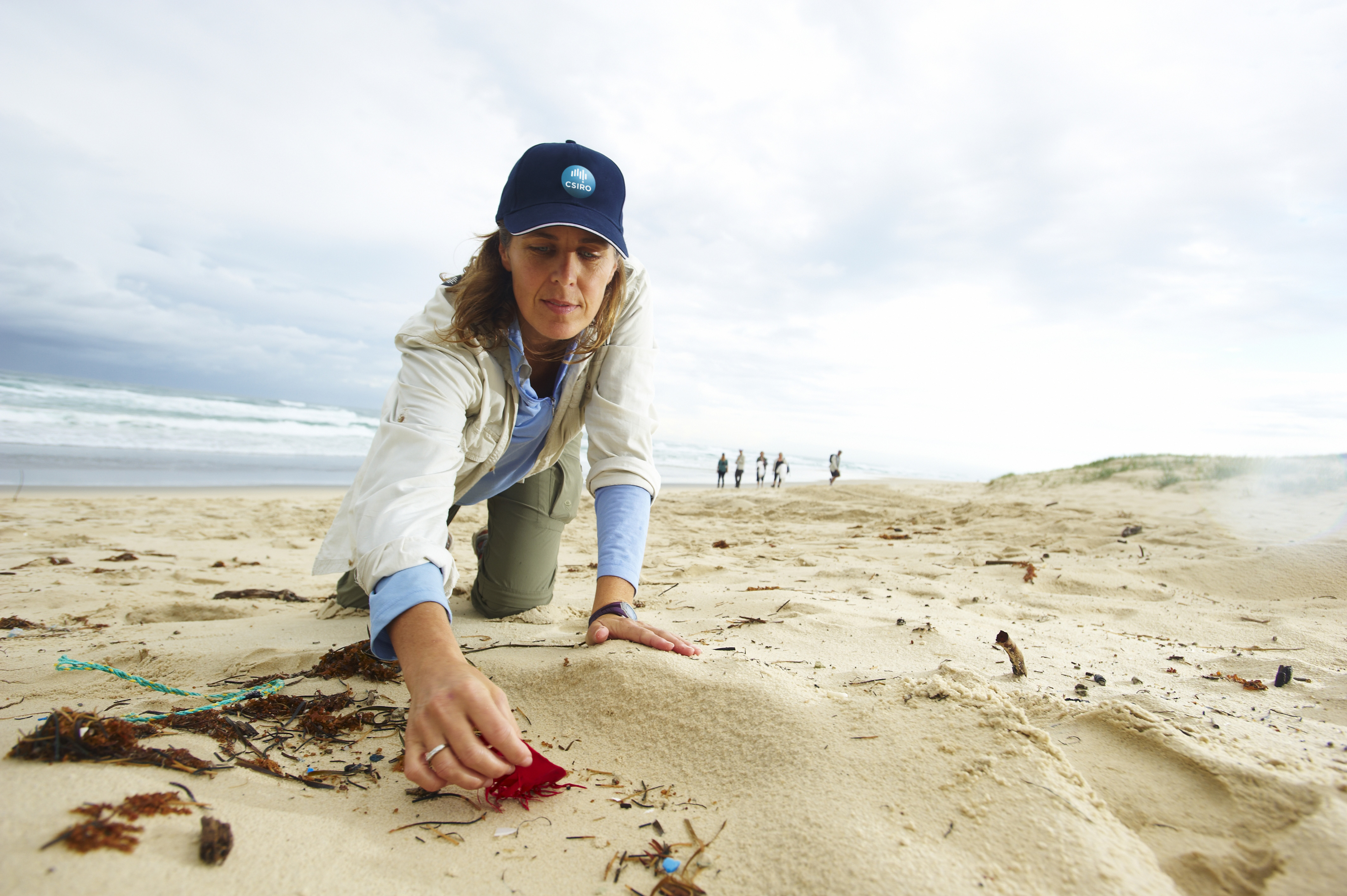 Researcher Dr Britta Denise Hardesty kneeling on a beach wearing a CSIRO cap, picking up a piece of a red balloon.