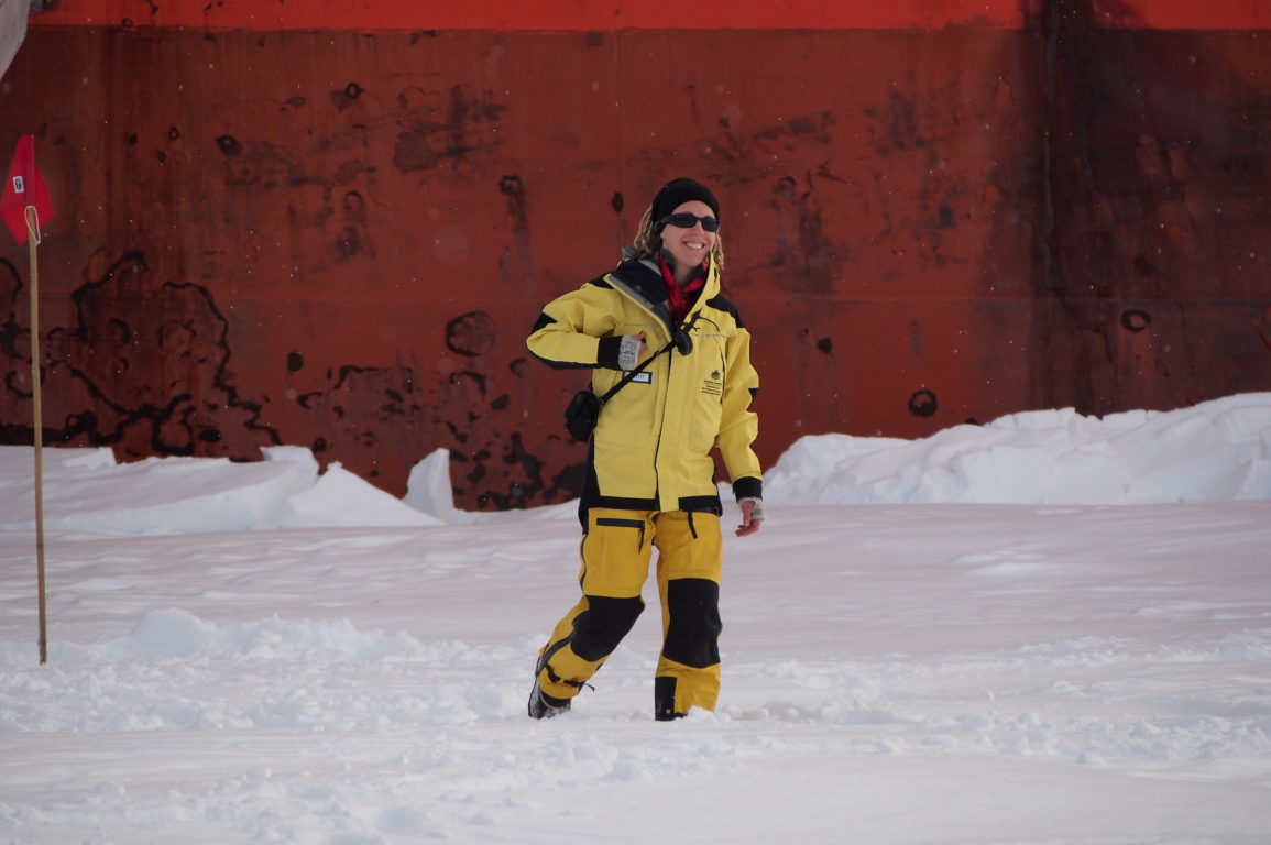 Jess Melbourne-Thomas undertaking sea ice ecosystem field work in East Antarctica. She wears a yellow suit and is surrounded by snow.