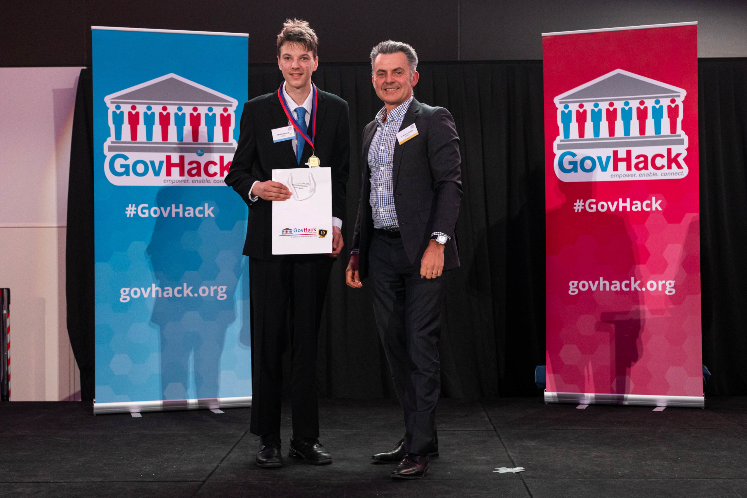 GovHack award winner holds a GovHack bag and is standing next to Director of the Atlas of Living Australia with GovHack banners in the background