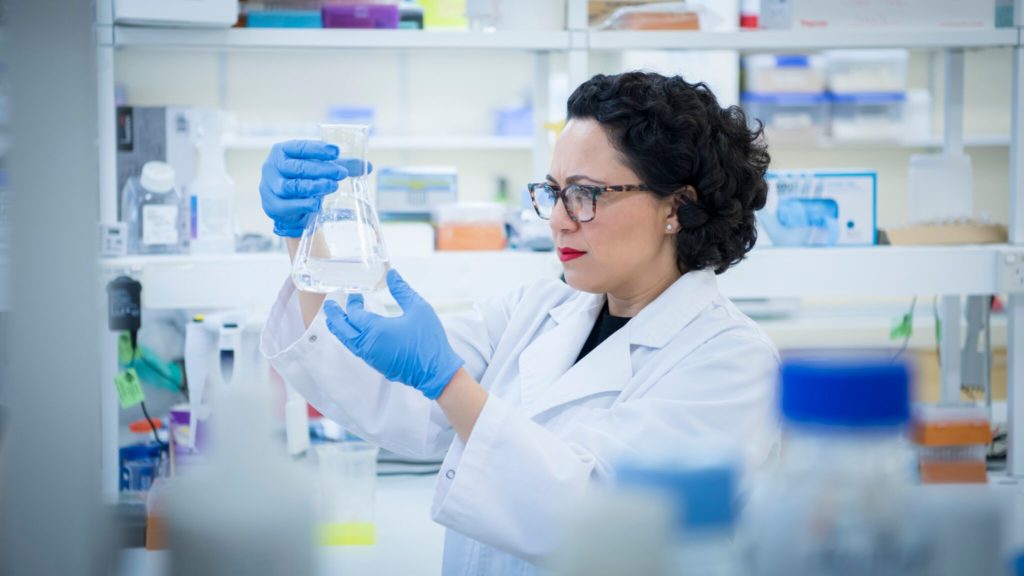 A woman in a white lab coat, glasses and blue gloves stares at a beaker of liquid.