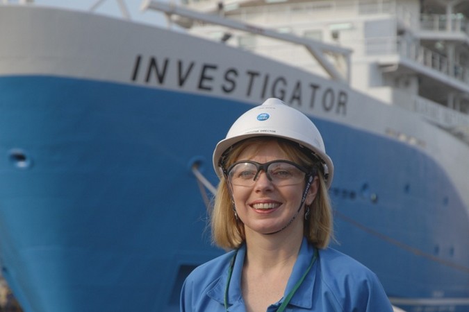 Toni Moate stands in front of docked research vessel wearing hardhat and safety glasses.
