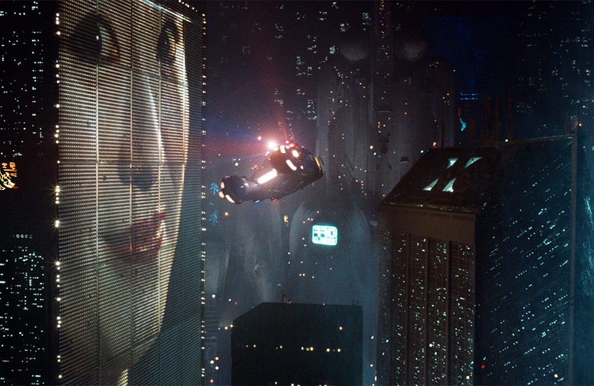 clip from the film Blade Runner showing flying cars around buildings at night. One building has a giant billboard of a lady's face