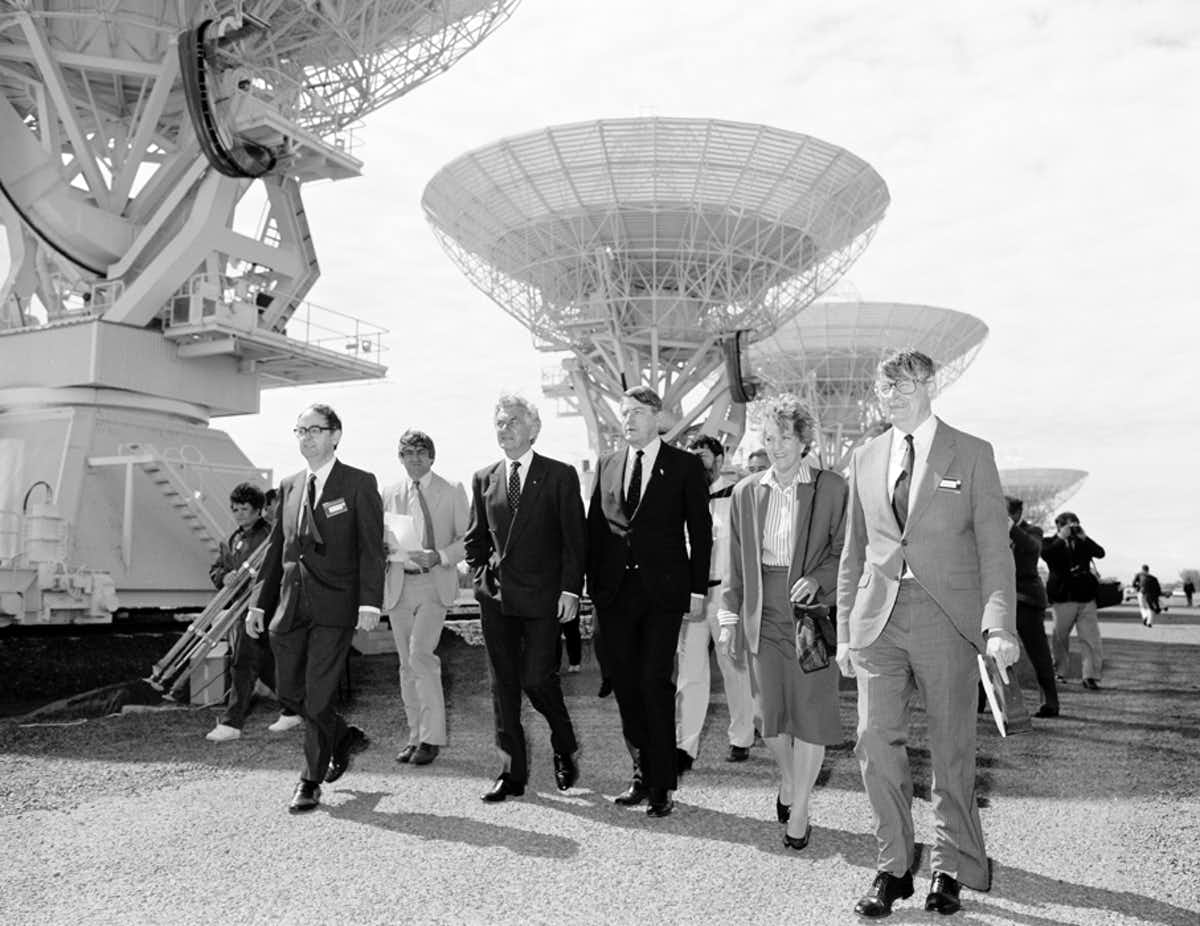 Black and white phot of Bob Hawke and a group of people walking next to telescope dishes