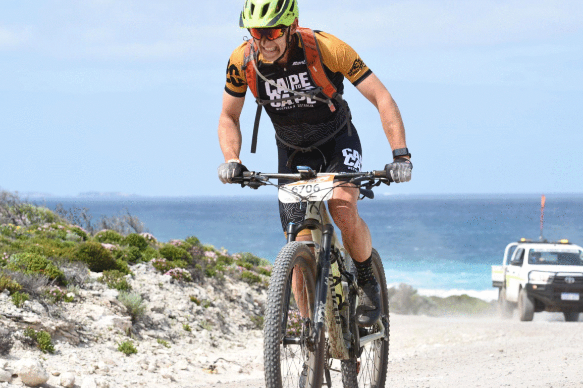 A man riding a bike on a dirt road with a 4WD and a beach behind him