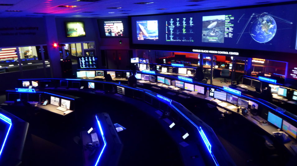 a circular control room with rows of desks, back lit in blue. Walls are covered in screens showing various images of earth and satellite dishes. Various technologies are lit up in the room.