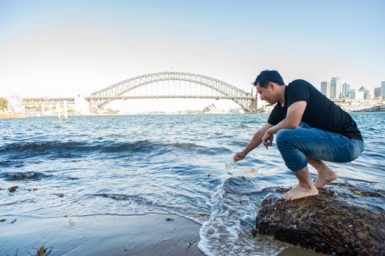 Man squatting down to collect water sample in front of Sydney Harbour Bridge