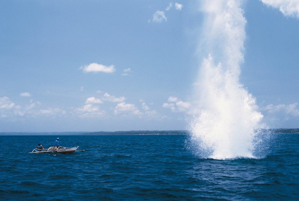 Fishing boat next to an explosive going off in the ocean, resulting in water going into the air