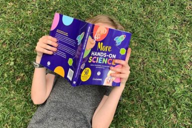 child laying on grass reading hands on science book