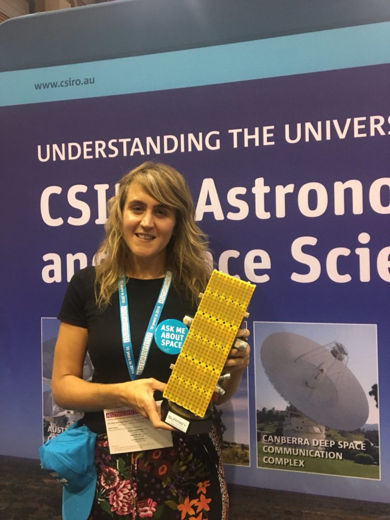 Dr Amy Parker smiling at the camera holding a prop in front of a space poster. She is wearing a lot of CSIRO merch