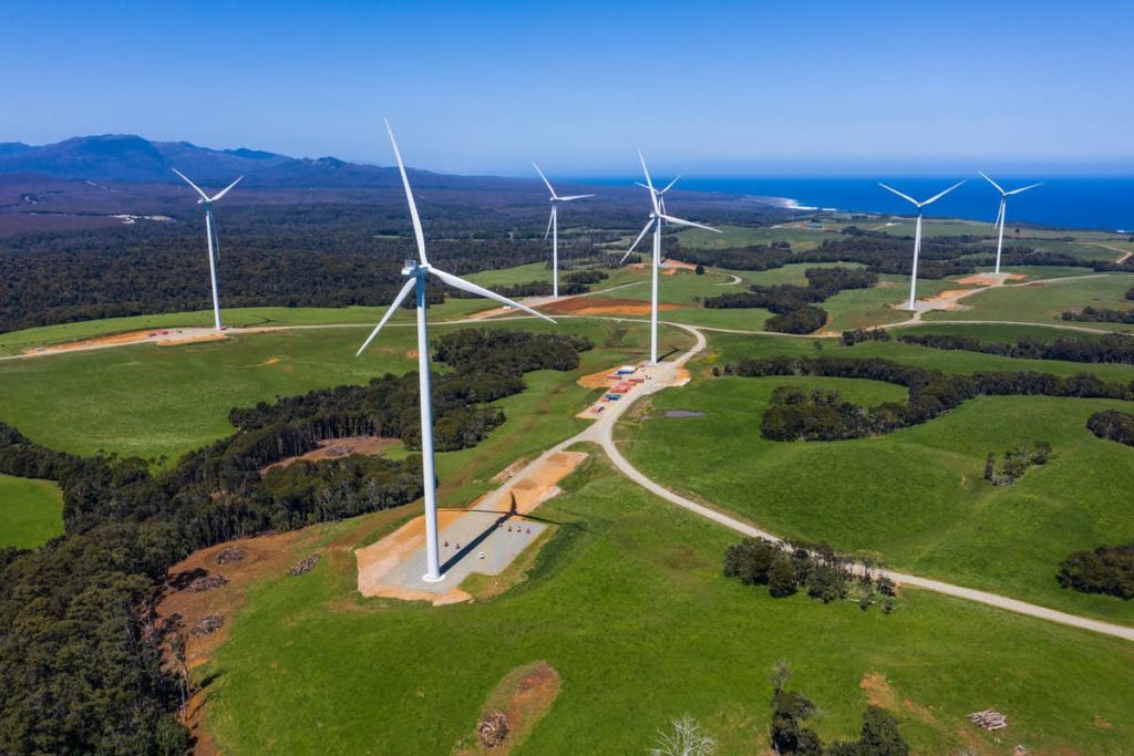 An aerial photo of wind turbines in a green farm area with the ocean in the background.
