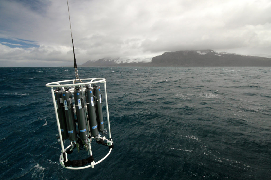 A piece of scientific equipment dangles above the ocean surface with an island in the background.