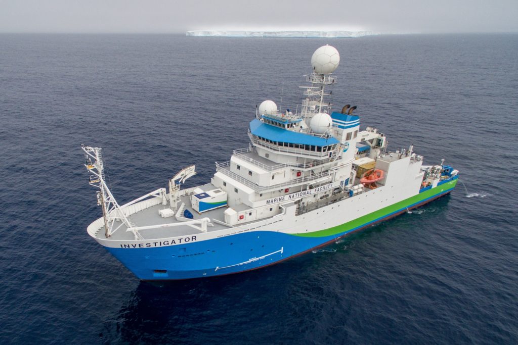 A blue, green and white research vessel with an iceberg in the background.