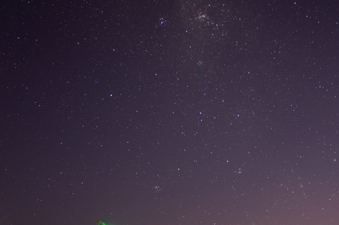 A photo taken at night shows a field of stars stretching across the sky above a few radio telescope dishes lit with green lights.