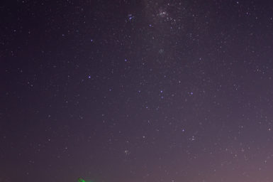 A photo taken at night shows a field of stars stretching across the sky above a few radio telescope dishes lit with green lights.