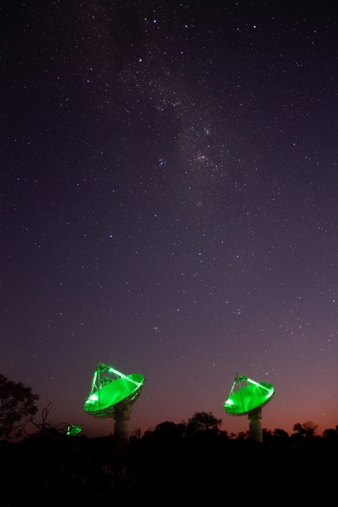 A photo taken at night shows a field of stars stretching across the sky above a few radio telescope dishes lit with green lights. 