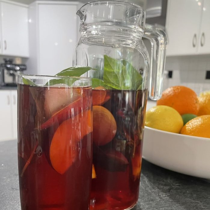 refreshing drinks in a glass jug and a tall grass. the drink is dark red with pieces of fruit and basil leaves