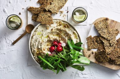 This healthy Christmas recipe is shown as an looking straight down on a bowl of dip surrounded by seed crackers and green leaves