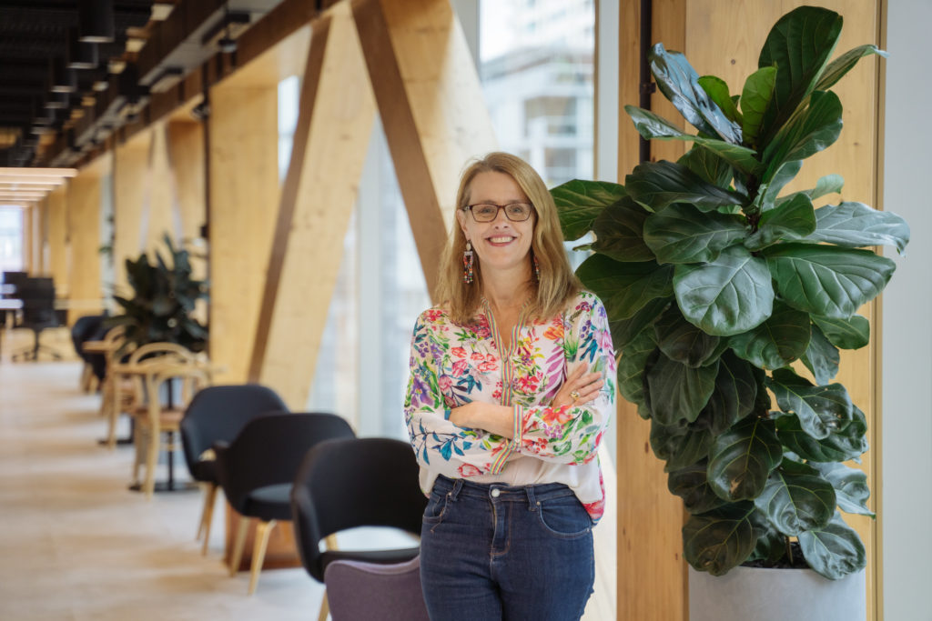 Pandemic pivot. HowToo co-founder Lisa Vincent wearing glasses, jeans and floral blouse in an open office with plants and black chairs. 