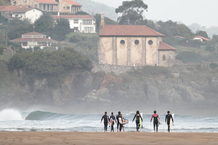 Surfers walking towards water with a rocky cliff and buildings above in background.